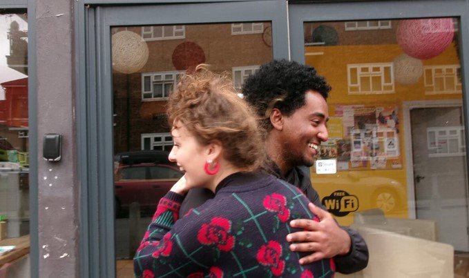The image shows two people embracing in front of a building with glass doors. The person on the left is wearing a sweater with a floral pattern, and the person on the right is smiling. The background includes a Free WiFi sign and some