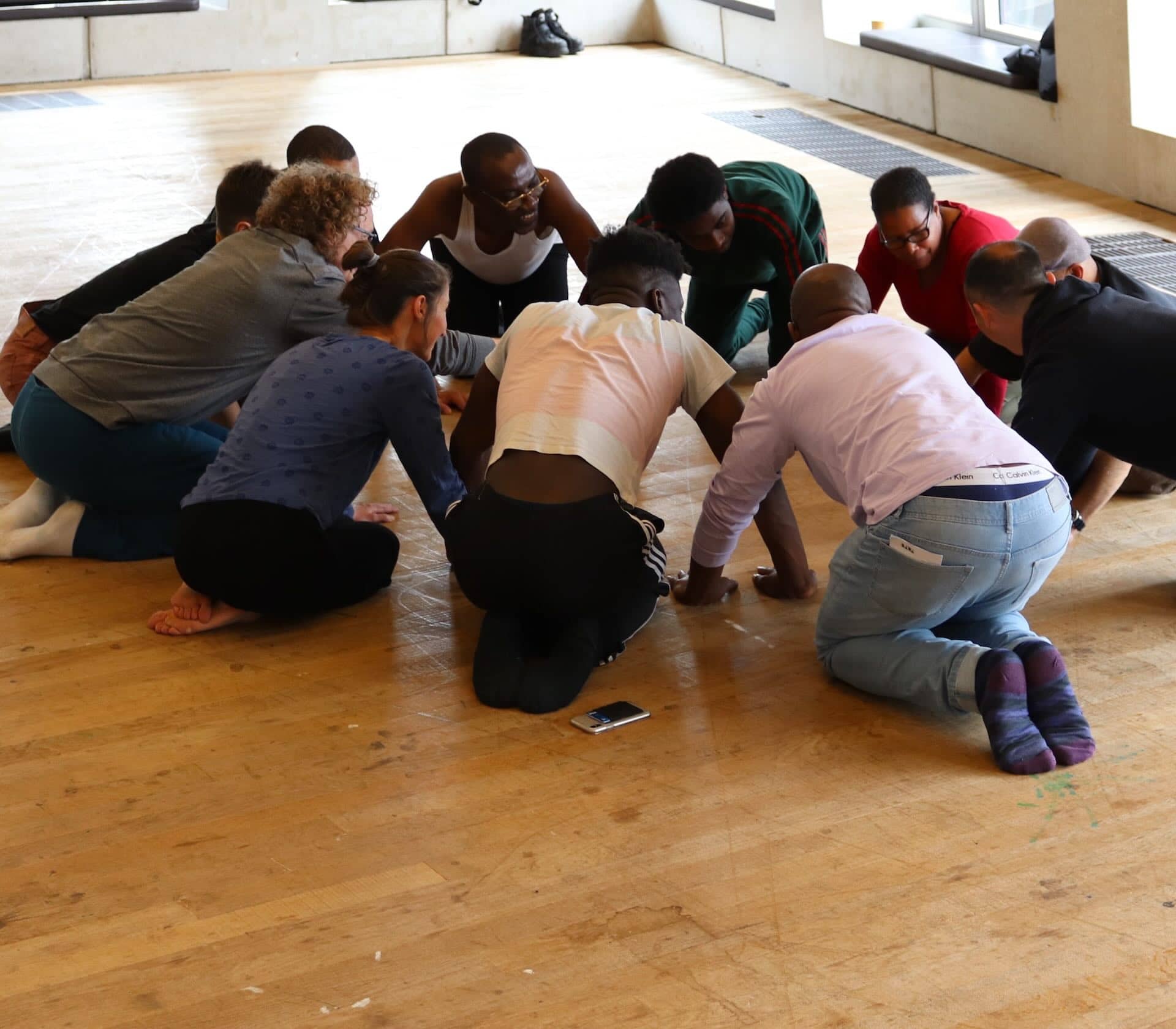 This image shows a group of people kneeling in a circle on a wooden floor. They appear to be engaged in an activity or discussion, with their heads close together. A smartphone is visible on the floor nearby. The setting looks like an indoor space