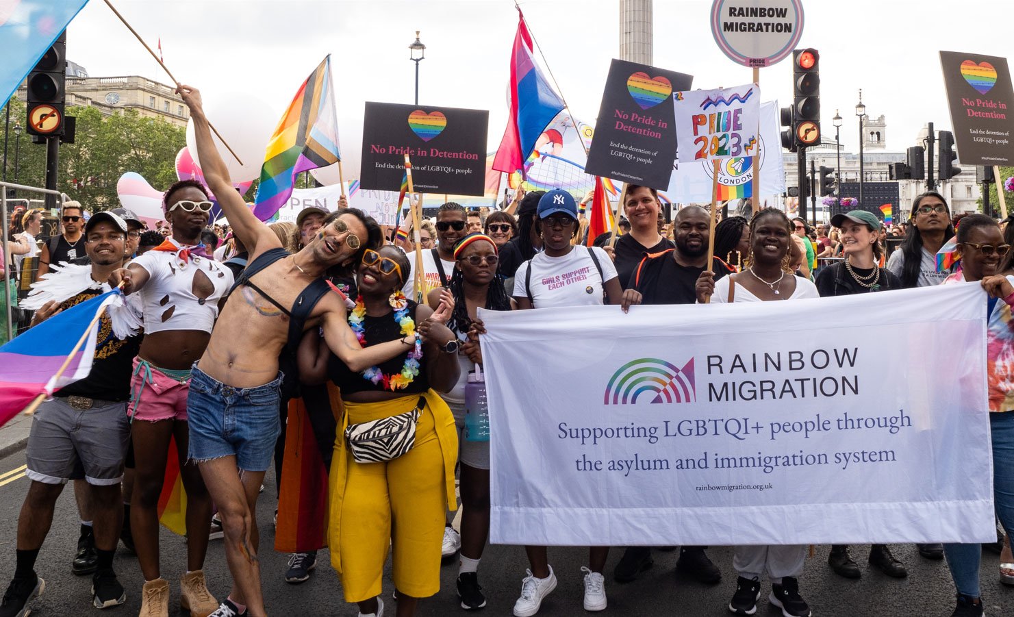 The image shows a group of people participating in a pride parade. They are holding a large banner for Rainbow Migration which supports LGBTQI+ people through the asylum and immigration system. Participants are carrying rainbow flags and signs with messages like No Pride
