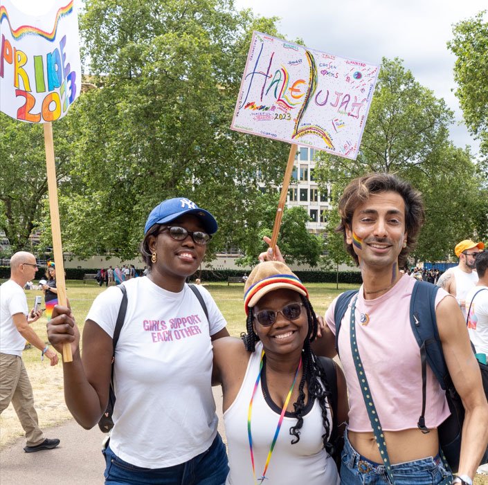 A group of young migrant people march in a pride parade, wearing rainbow face paint and holding Pride signs
