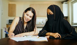 The image shows two women sitting at a table. One woman is wearing a striped sleeveless top, holding a pen, and appears to be smiling while looking at some papers or a magazine. The other woman is wearing a black niqab and is looking