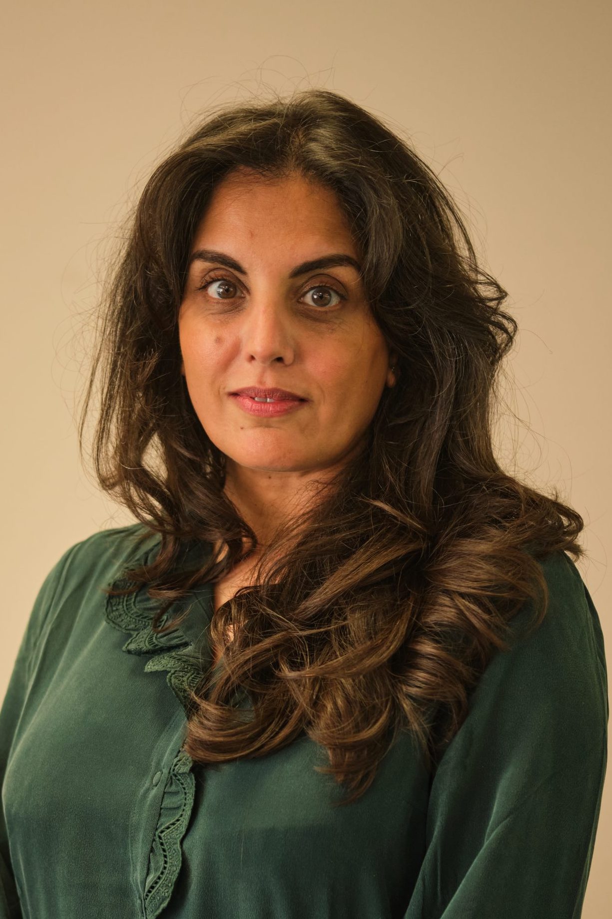 The image is a portrait of a person with long, dark hair and a serious expression. They are wearing a dark green shirt and are looking directly at the camera against a neutral background.