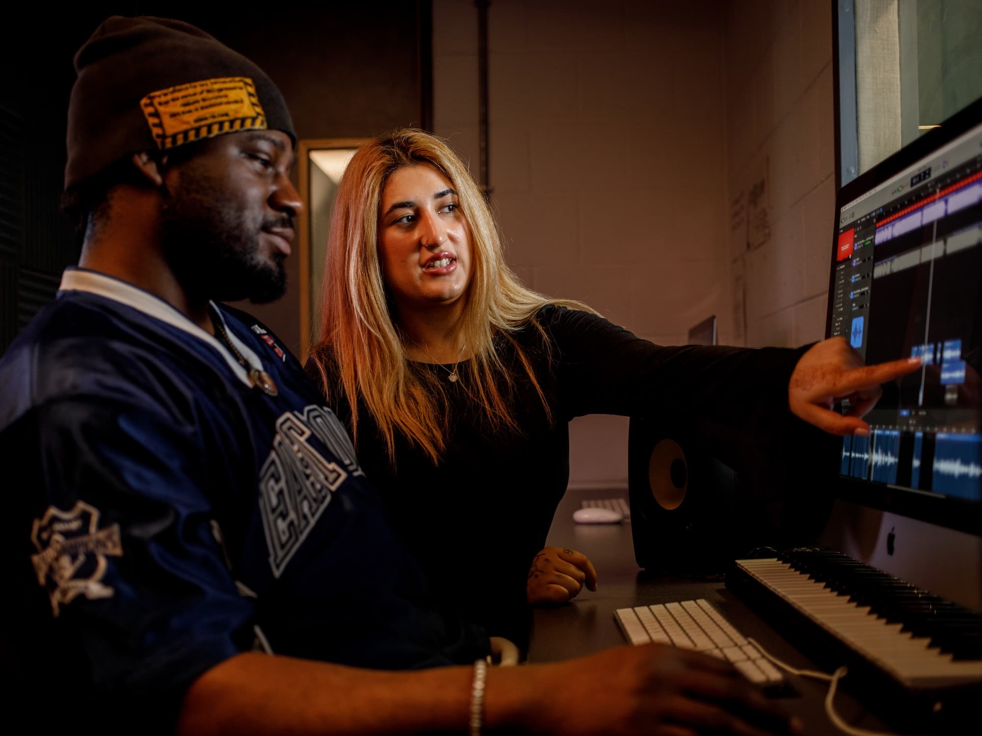The image shows two people in a studio setting. One person is seated at a computer with a keyboard in front of them, looking at the screen, while the other person is standing and pointing at the monitor. The screen displays what appears to be audio
