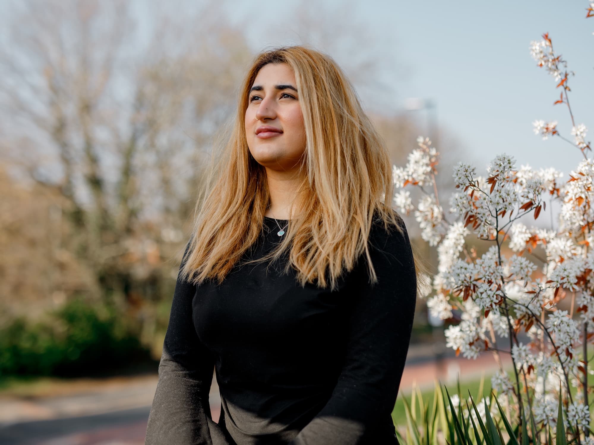 The image shows a person with long hair standing outdoors near blooming white flowers. They are wearing a black top and the background includes trees and a clear sky. The person is looking slightly upwards.