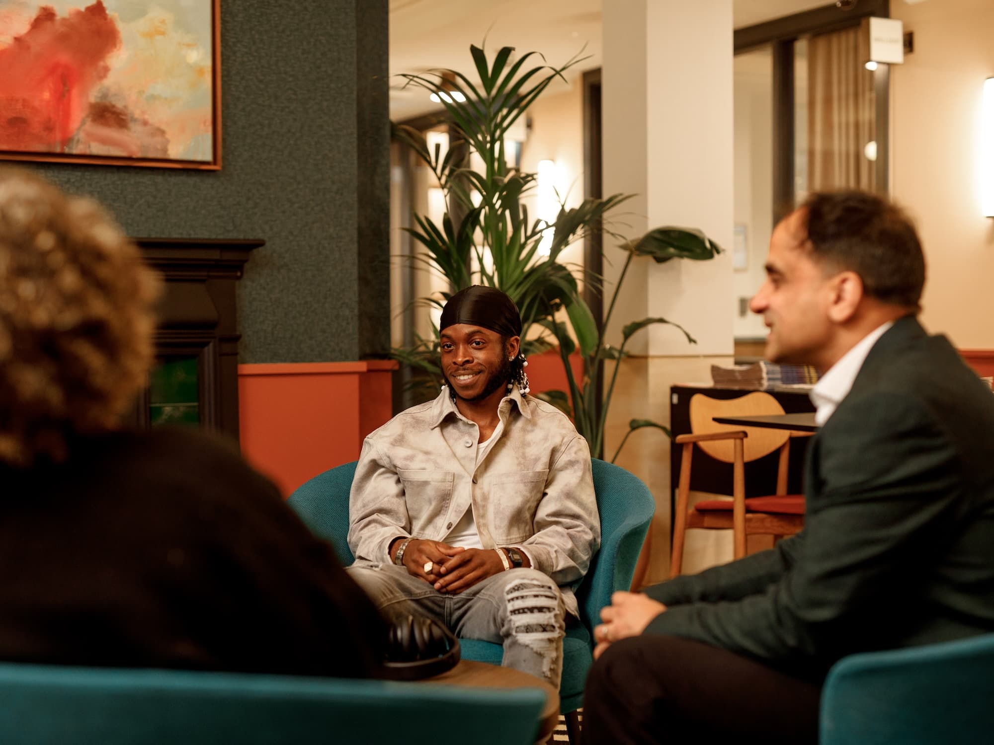 The image shows three people sitting in a cozy indoor setting. They appear to be engaged in a conversation. There is abstract artwork on the wall, a large plant in the background, and modern furnishings, including chairs and a table. The atmosphere seems relaxed