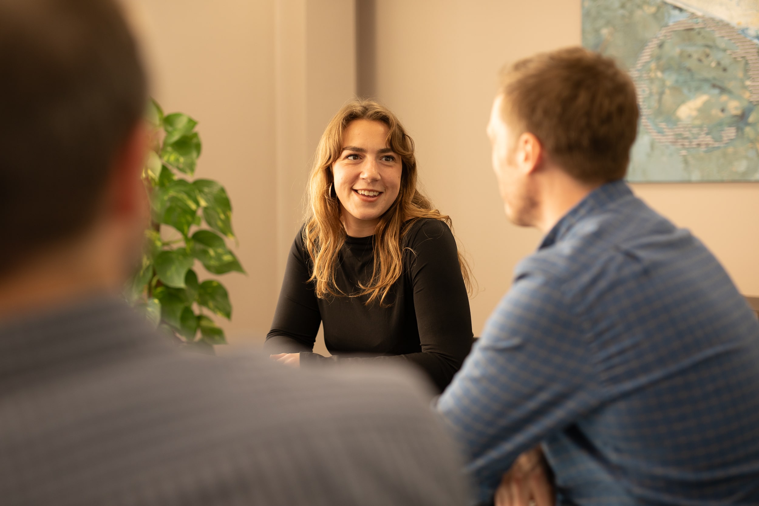 The image shows three people in a casual indoor setting having a conversation. One person, facing the camera, is smiling, while the other two have their backs to the camera. There is a plant in the background and a piece of artwork on the wall