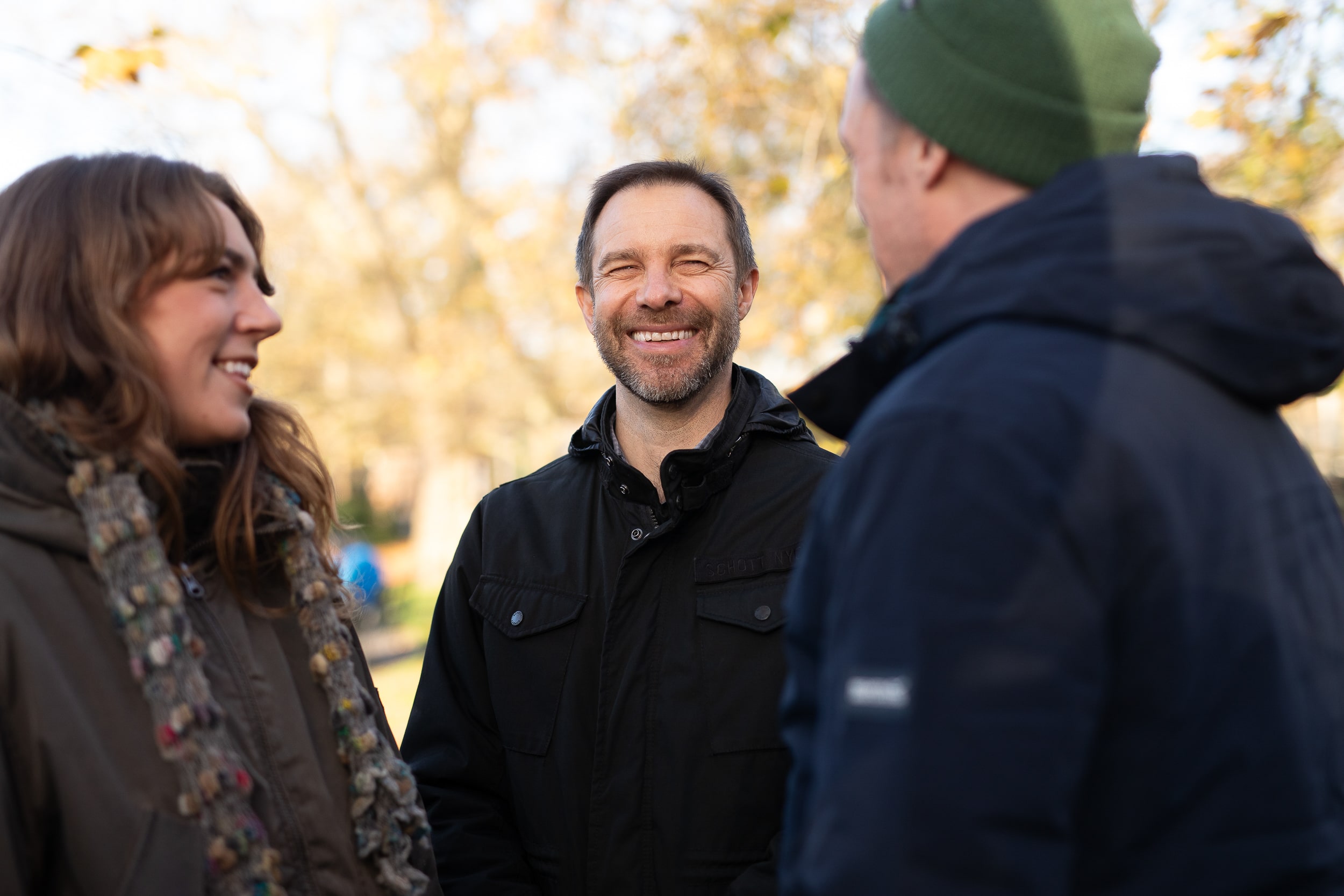 The image shows three people outdoors, engaging in a conversation and smiling. They are wearing winter clothing, including jackets and a beanie. The background is blurred, suggesting they are in a park or a natural setting.