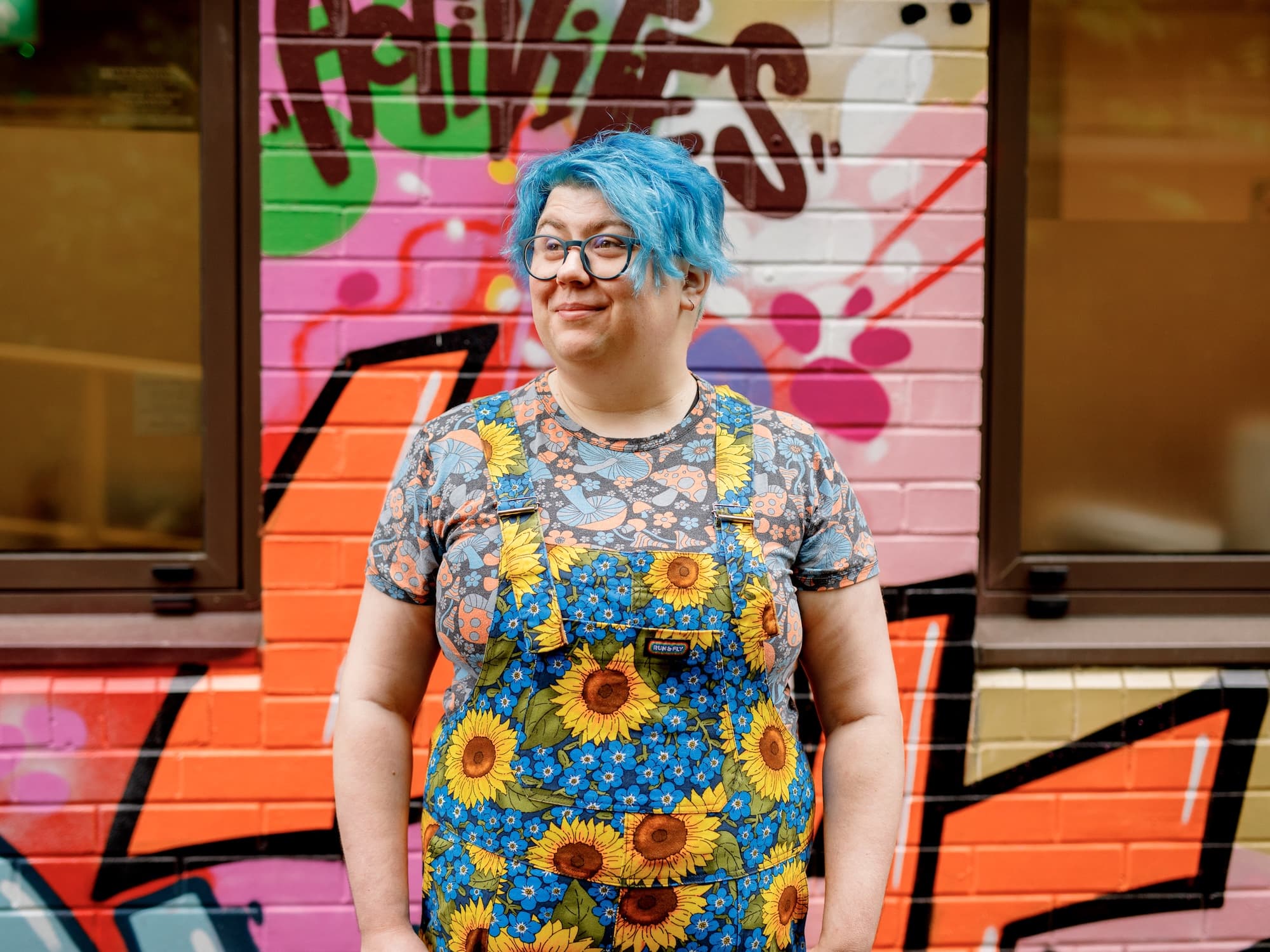 The image shows a person with blue hair standing in front of a colorful graffiti-covered wall. The person is wearing glasses, and their outfit features sunflowers and floral patterns. The background is vibrant, with various colors and artistic designs.