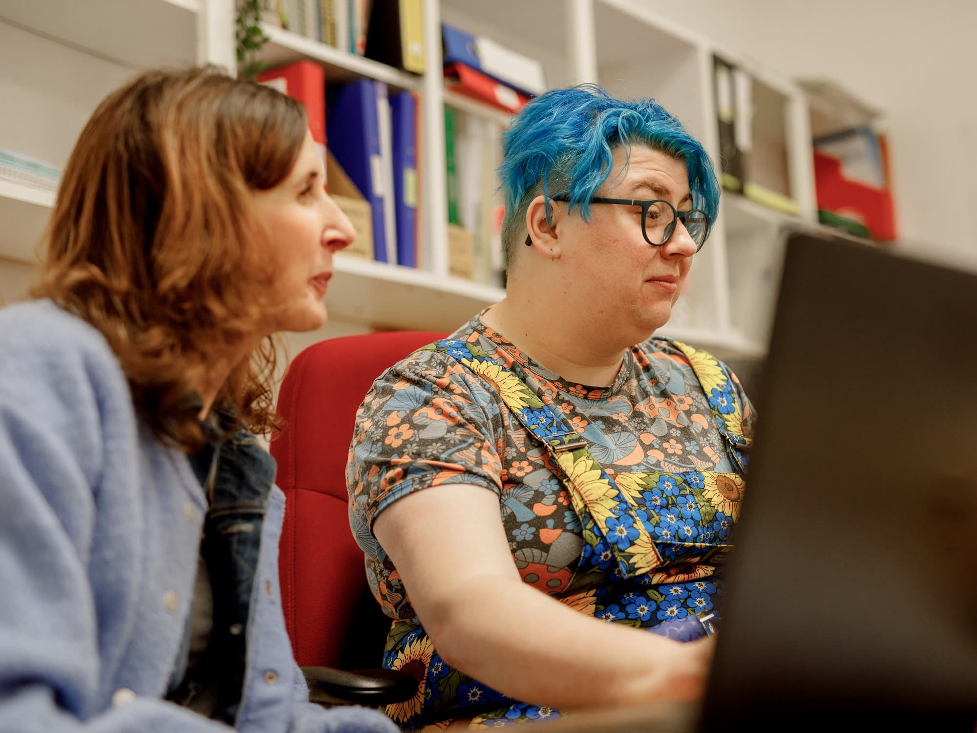 The image shows two people sitting together, possibly working or discussing something while looking at a laptop. They are in an indoor setting with shelves containing books and binders in the background. The person on the right has bright blue hair and glasses, and is