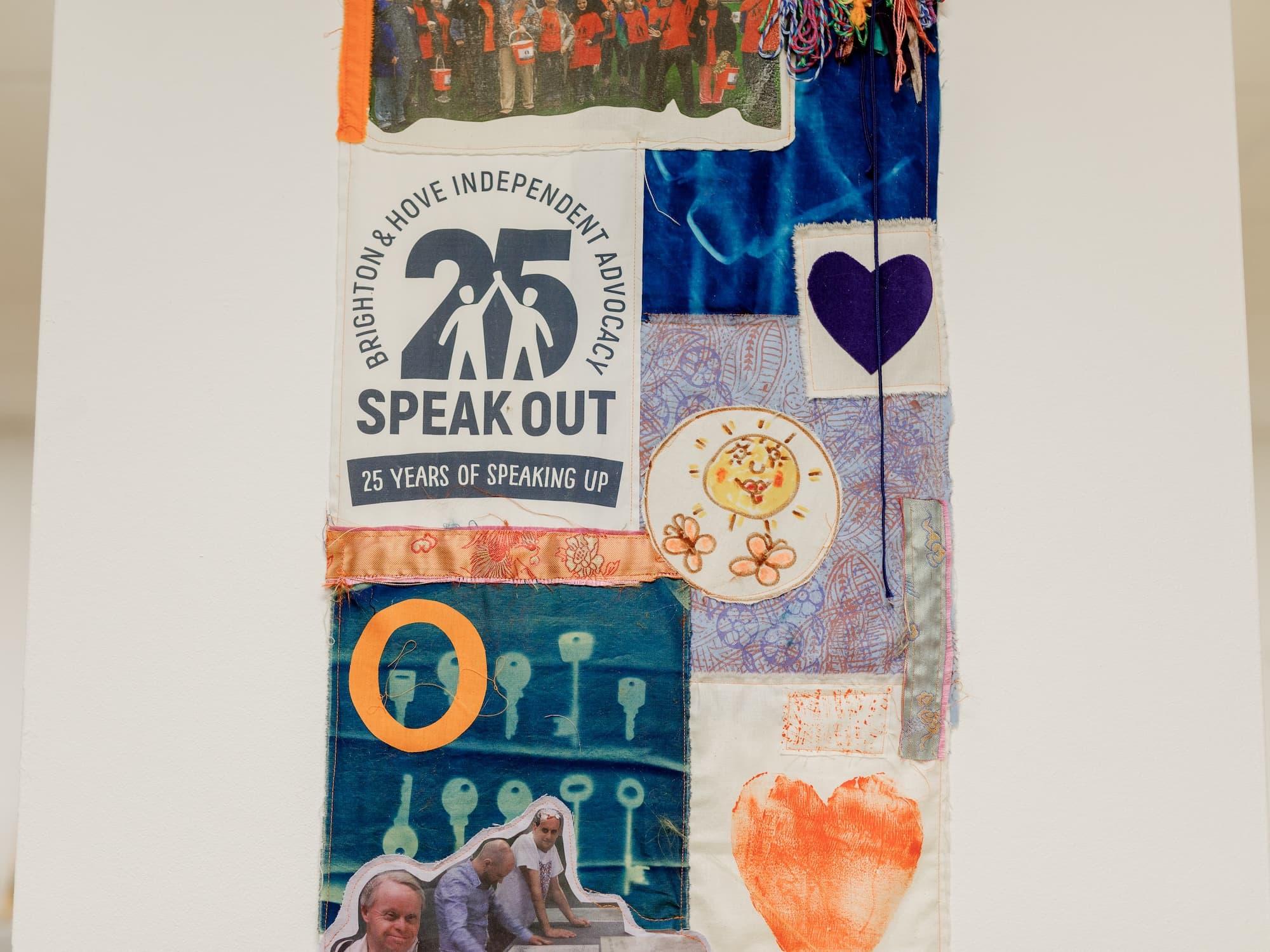 This image features a colorful textile or quilt-like artwork. It includes various sections with different designs, such as: - A section with the text Brighton & Hove Independent Advocacy 25 Speak Out - 25 Years of Speaking Up. - A