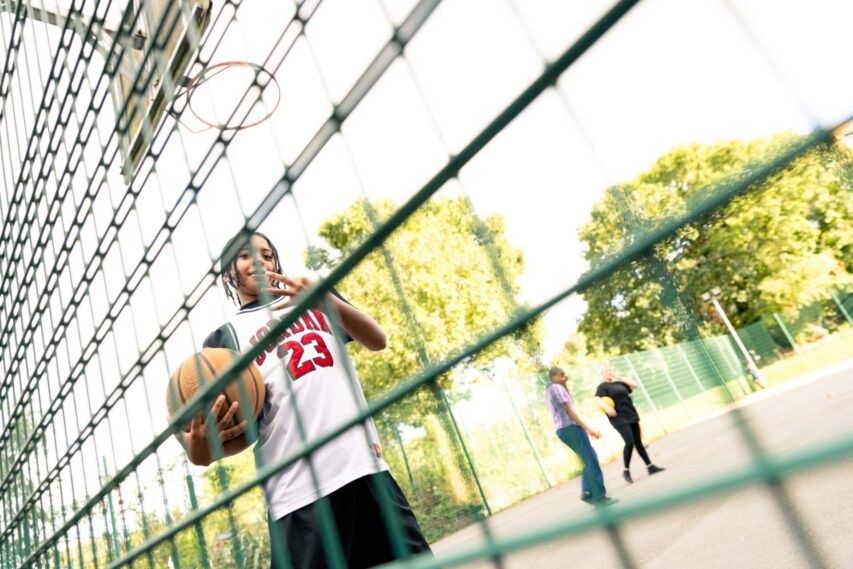 A young person holds a basketball and looks through a green fence on a basketball court surrounded by trees