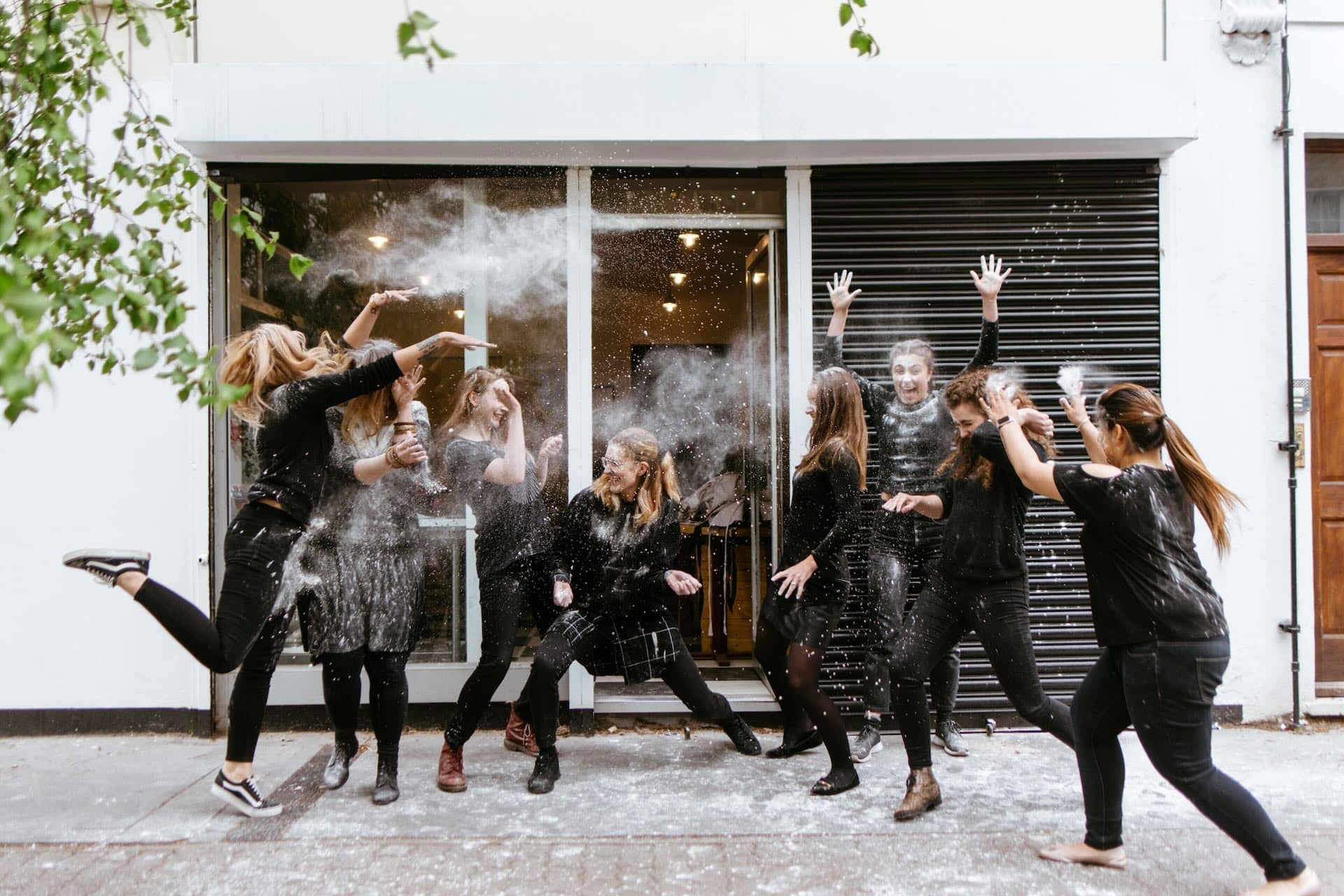 The image shows a group of people having fun and throwing what seems to be powder or flour at each other. They are outside in front of a building, with sliding doors and a window behind them. Everyone appears to be wearing casual clothing, and they