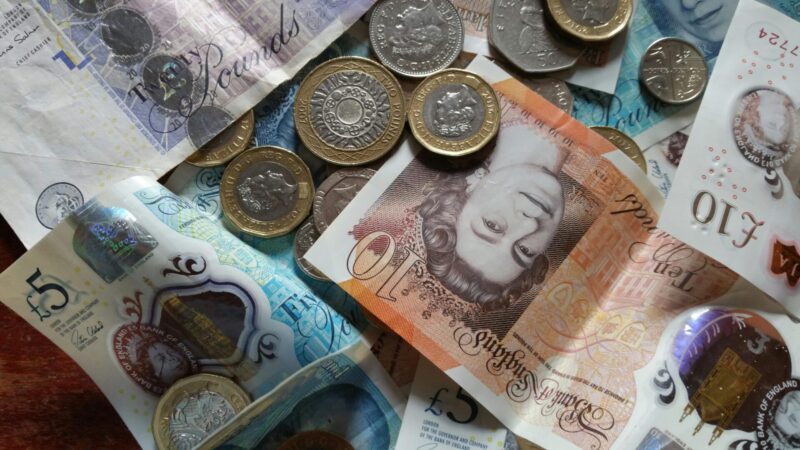 The image shows a collection of British currency, including banknotes and coins. There are £5 and £10 notes, as well as various coins scattered around.