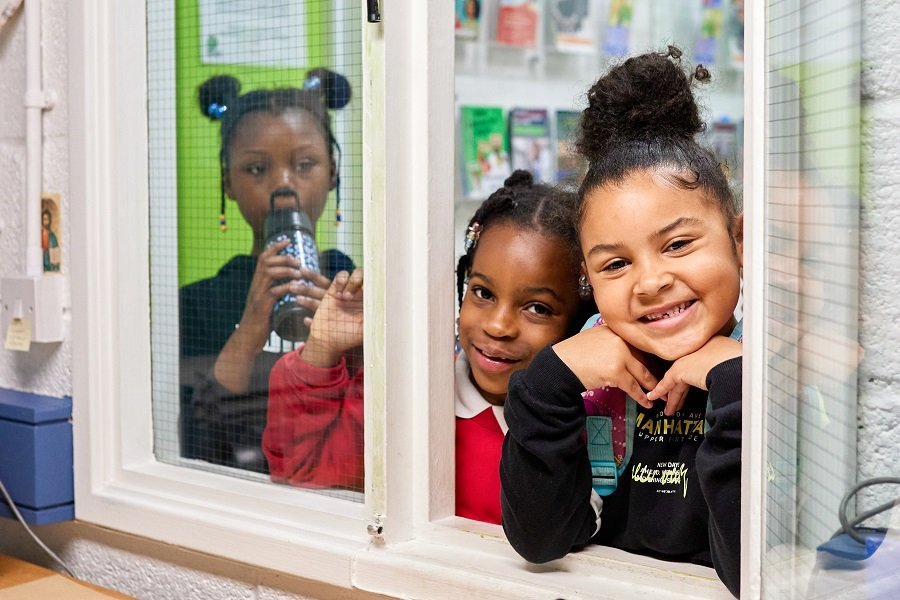 The image features three children. Two are smiling and looking through an open window or partition, while one is drinking from a bottle behind the glass. They appear to be in a classroom or educational setting, with posters or materials visible in the background.