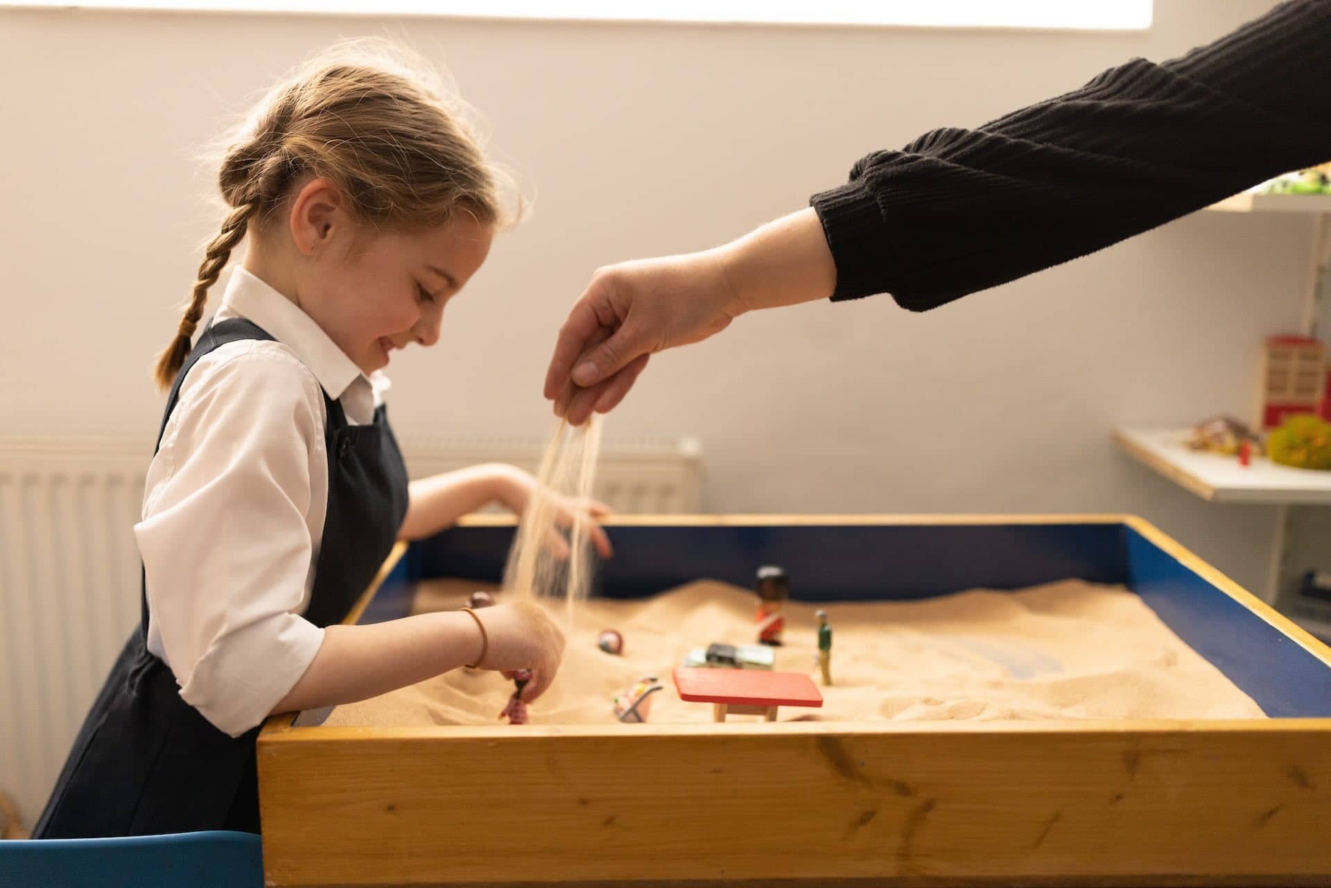 The image shows a young girl playing with a sand tray. She seems happy and engaged, using her hands to move objects around in the sand. Another person's hand is visible, pouring sand into the tray. There are small objects scattered in the tray,