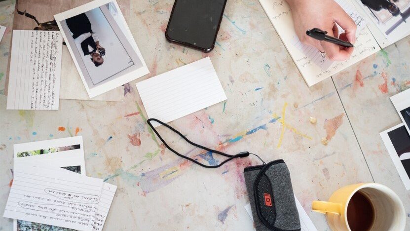 The image shows a cluttered workspace. There are several photographs and index cards scattered on a table. A person is writing on a piece of paper with a pen. A smartphone, a fabric case with a strap, and a mug with a drink are