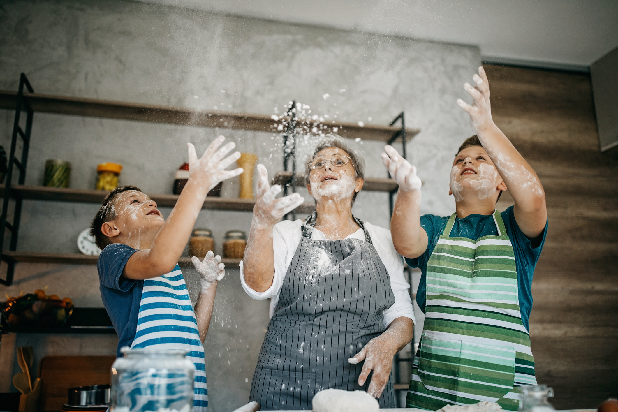 The image shows a group of people having fun and throwing what seems to be powder or flour at each other. They are outside in front of a building, with sliding doors and a window behind them. Everyone appears to be wearing casual clothing, and they