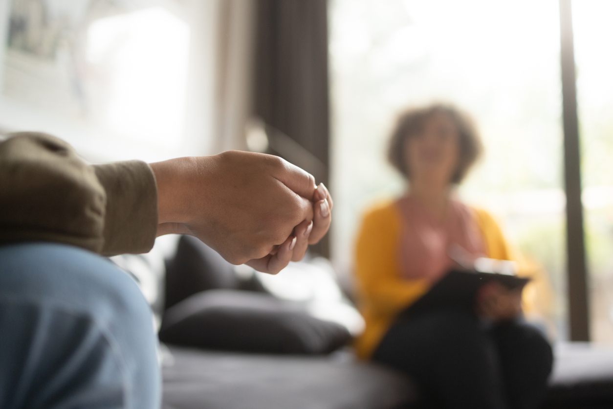 The image shows two individuals in a room. One person is visible in the foreground, with their hands clasped. In the background, there's another person sitting on a couch, holding a notepad. The setting suggests a conversation or therapy session.