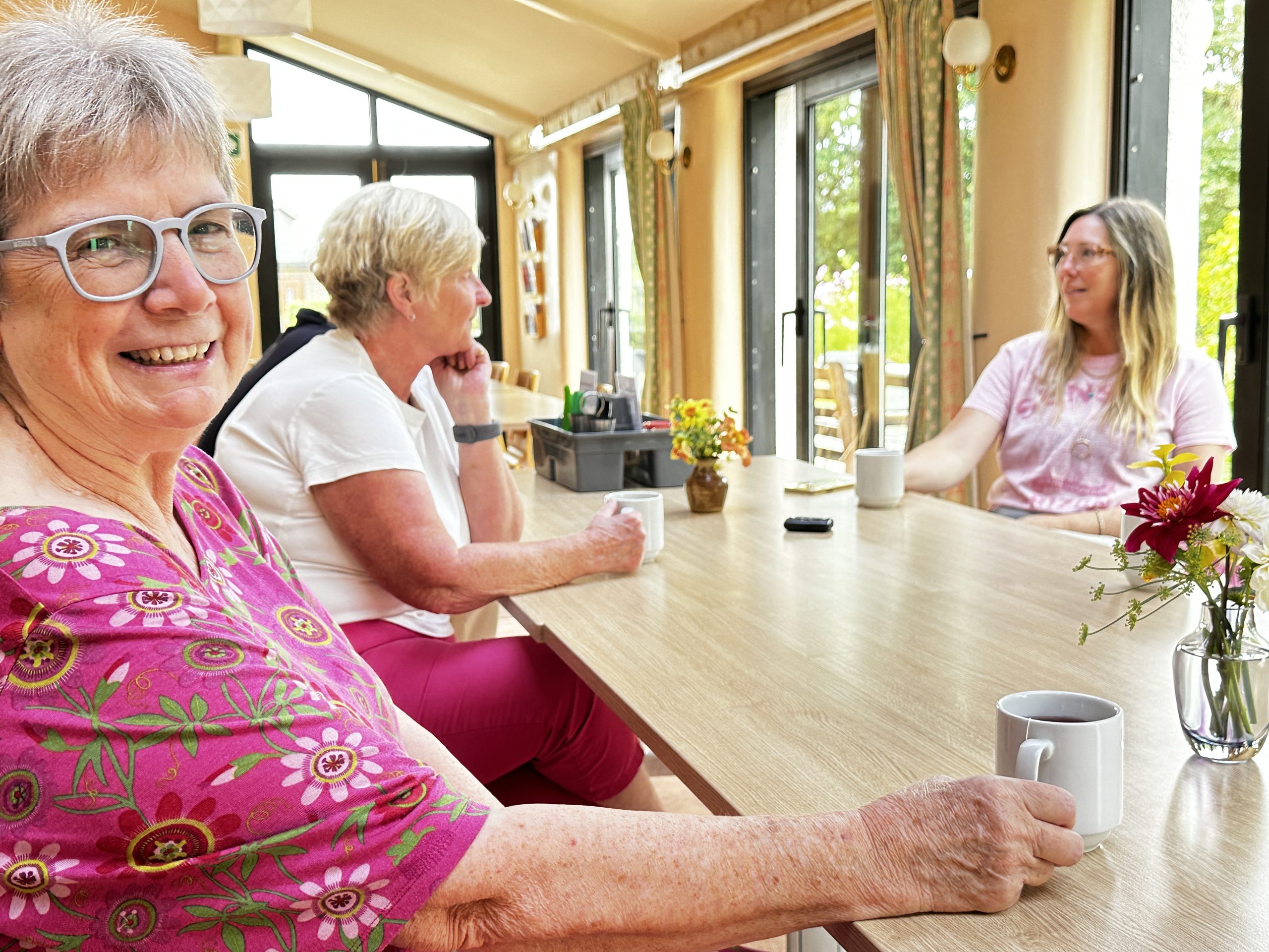 A woman in a pink top with a cup of tea smiles as she sits at a table near a window.