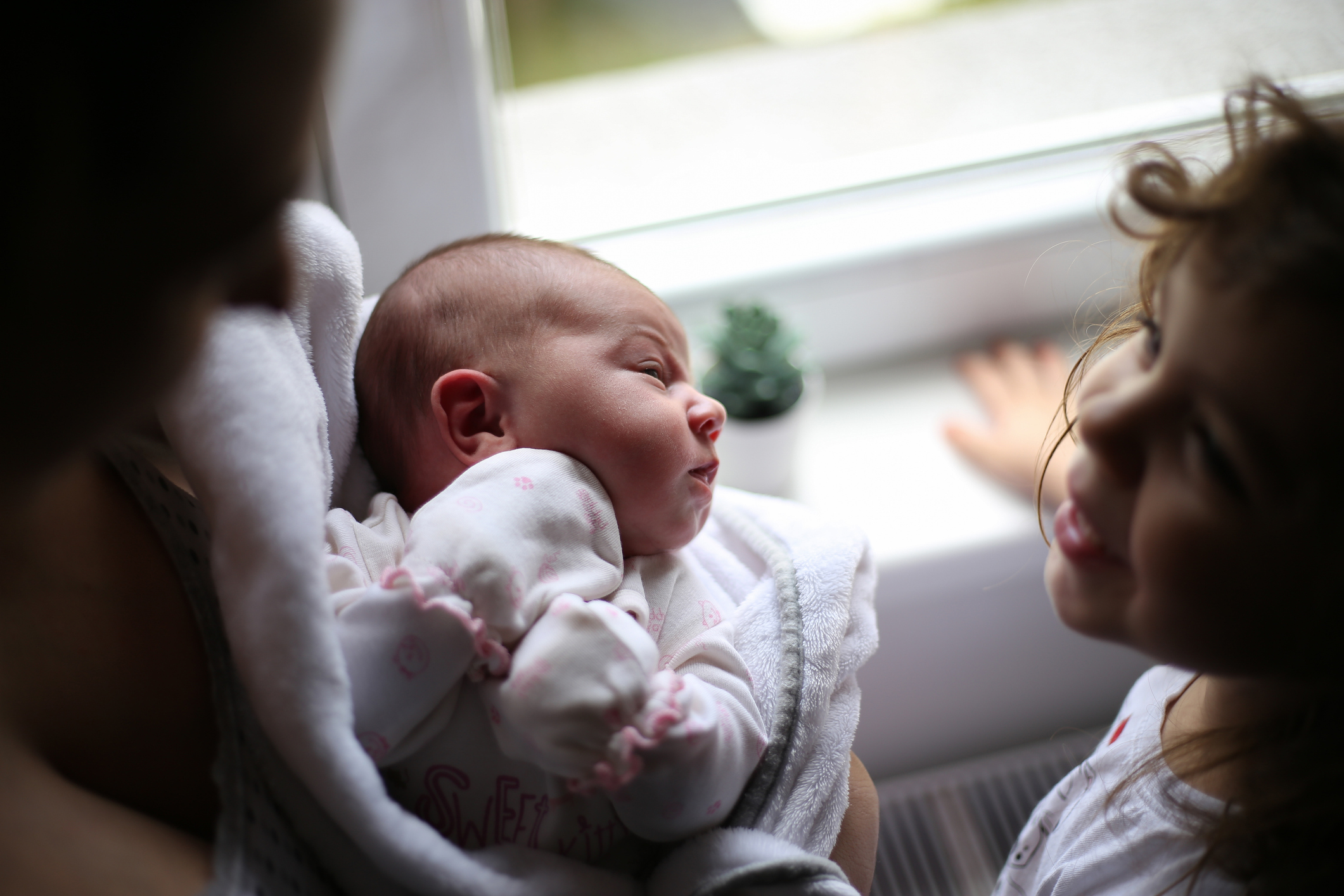 A parent holds a baby by a window so another, older child can meet them