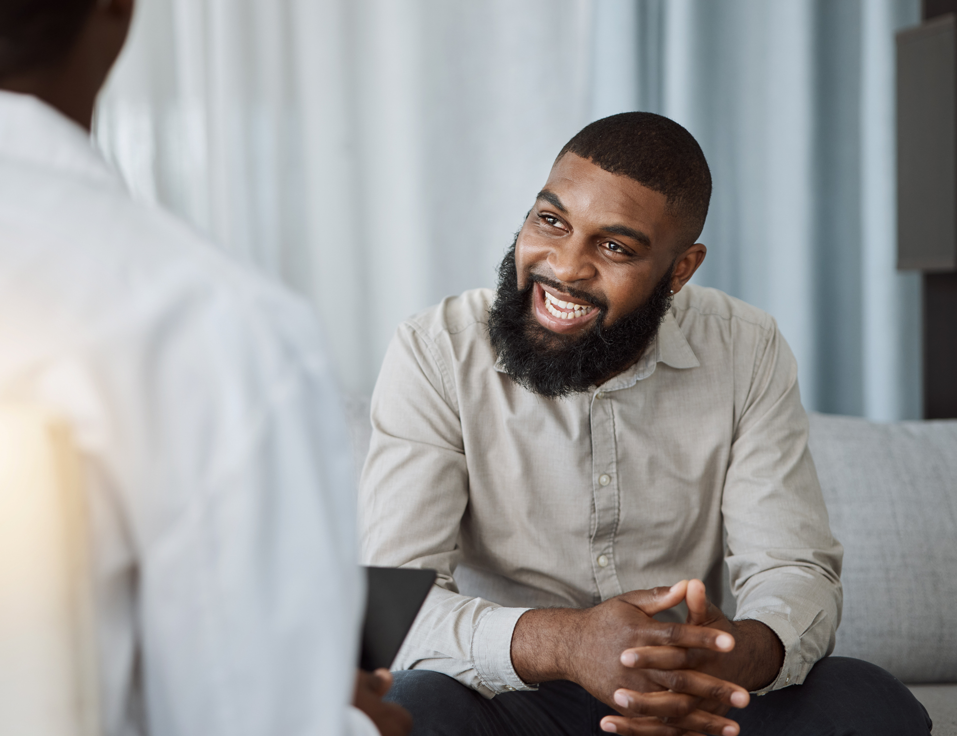 A smiling man sits on a sofa during a conversation with another person, who is seen from behind.