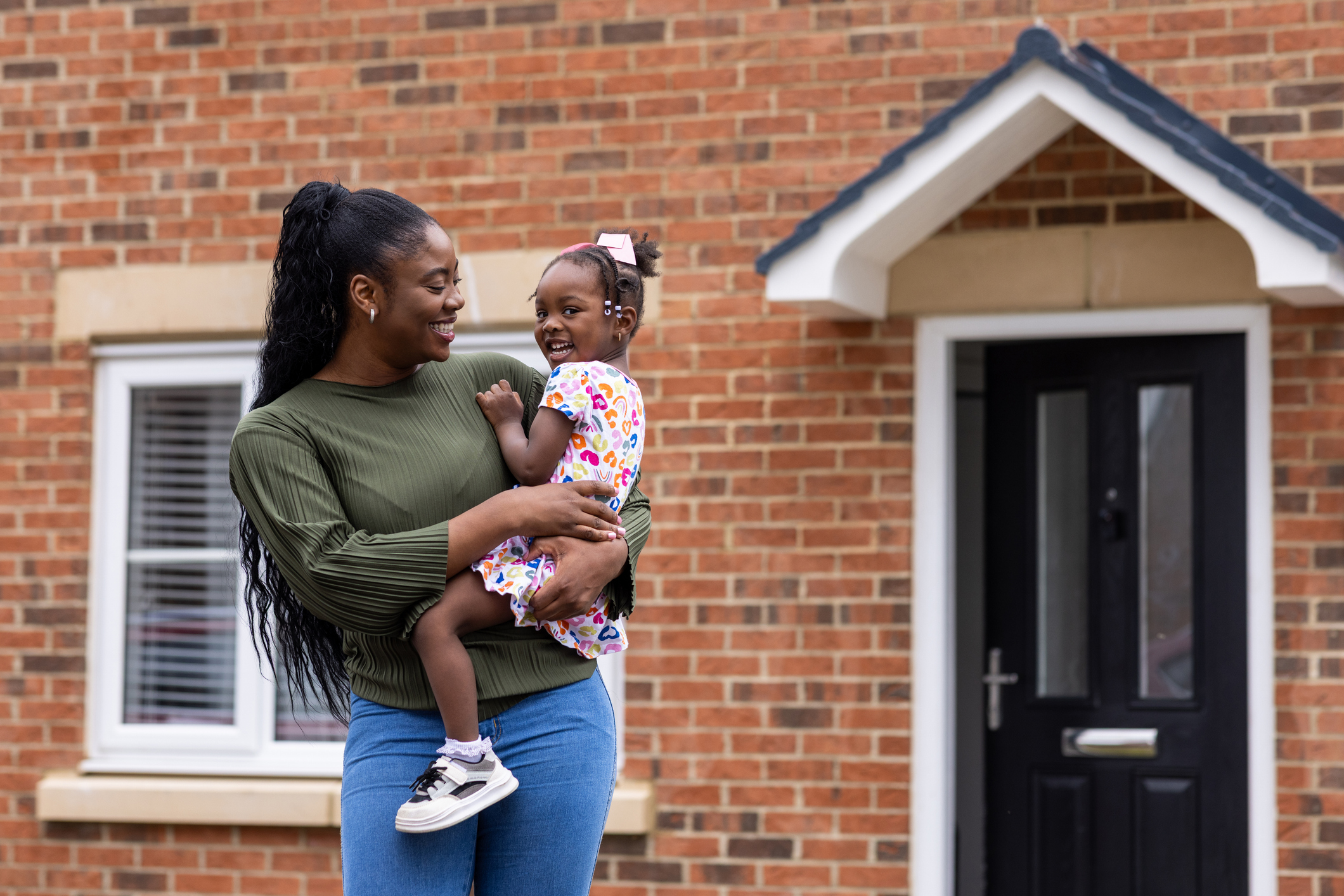 A smiling woman holds a young child outside a brick house with a black front door.