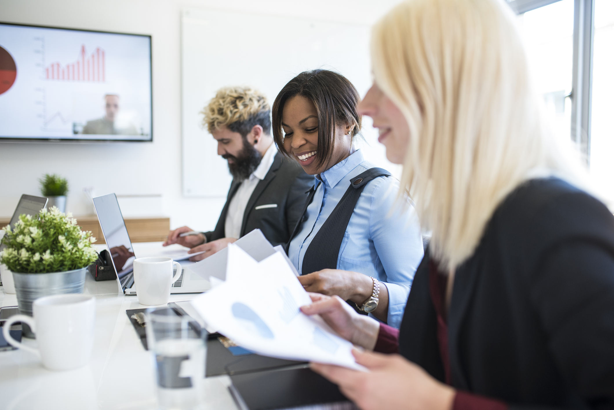 A group of colleagues sit around a meeting table reviewing printed documents, with one person smiling while others work on laptops and papers.