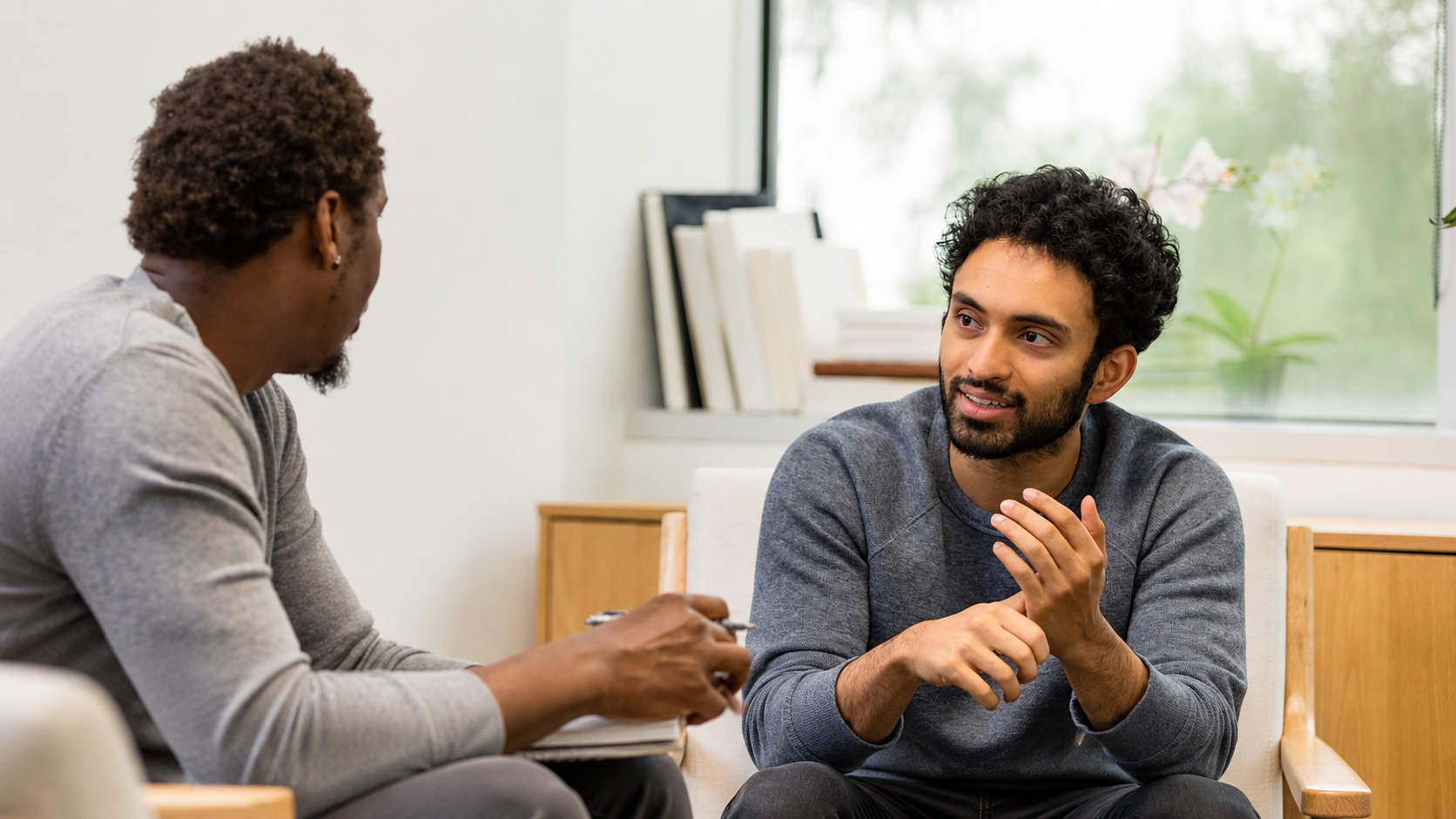 Two men sitting indoors in conversation, with one man taking notes while the other speaks.