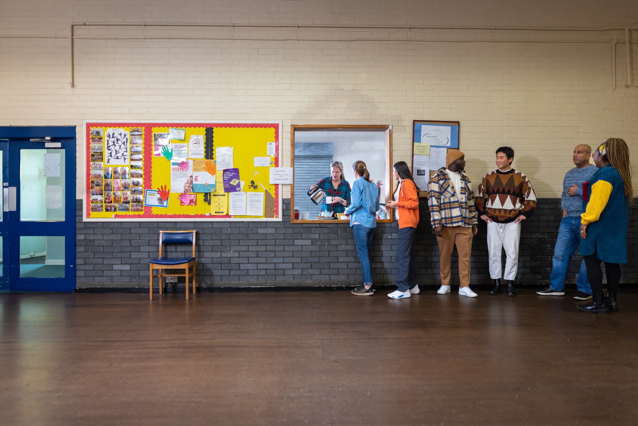 A group of people chatting in a community hall, with one person serving drinks near a noticeboard.