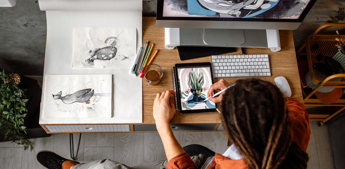 Illustrator working at a desk, drawing on a tablet with paper sketches alongside.