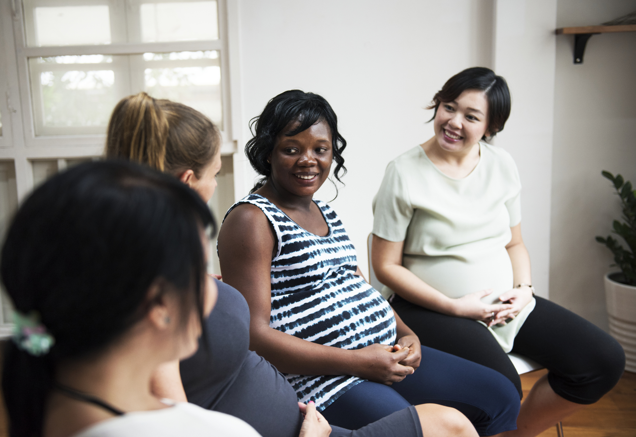 A group of pregnant women sitting in a circle, smiling and talking together in a bright room.